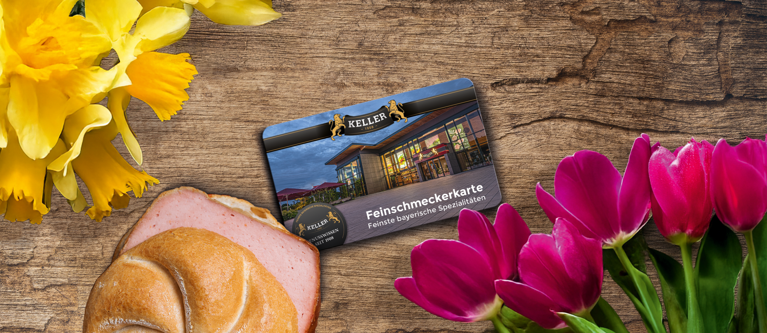 Feinschmeckerkarte auf Holztisch neben Leberkäs-Semmel, rosa Tulpen und gelben Narzissen.