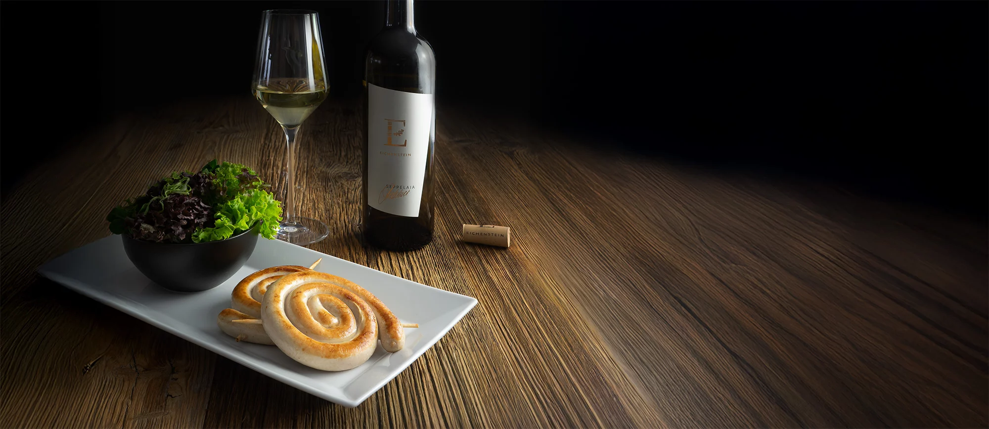 Edle Food-Fotografie auf dunklem Holztisch mit dramatischer Beleuchtung. Im Vordergrund eine weiße rechteckige Platte mit zwei spiralförmig aufgerollten Bratwürsten in goldbrauner Farbe. Links davon eine schwarze Schale mit frischem Blattsalat-Mix. Dahinter eine Weinflasche mit minimalistischem weißem Etikett und kupferfarbenen Details sowie ein gefülltes Weißweinglas. Rechts liegt ein Weinkorken. Dunkler, fast schwarzer Hintergrund mit gezieltem Spotlicht erzeugt Premium-Atmosphäre und betont die kulinarische Inszenierung. Hochwertige Produktfotografie im Fine-Dining-Stil.